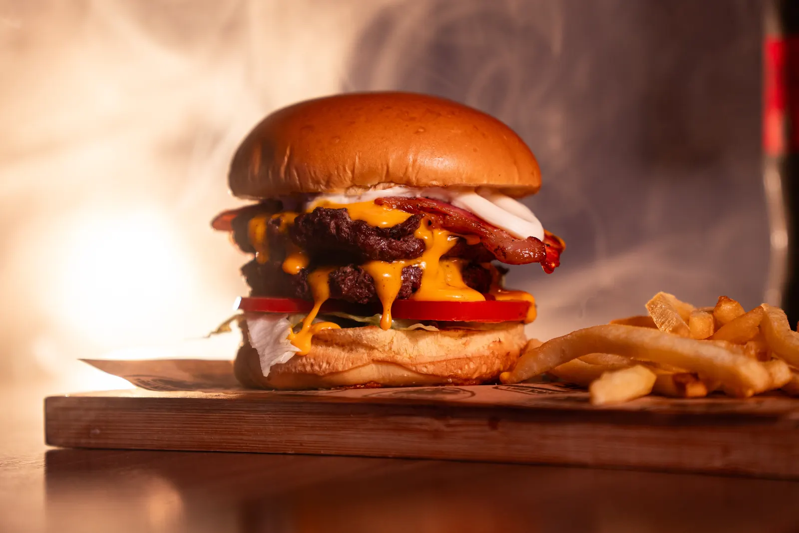 Hambúrguer duplo com cheddar derretido escorrendo, bacon e fumaça ao fundo, sobre tábua de madeira com batatas fritas
