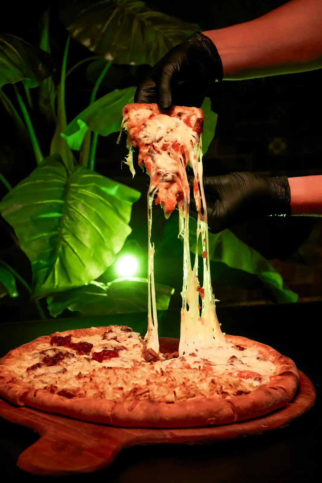Fatia de pizza sendo levantada com queijo derretido esticando, iluminação dramática verde ao fundo